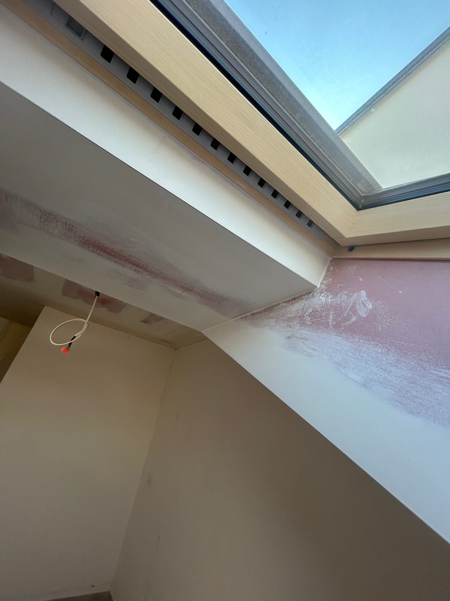 Ściana pod skosem z oknem dachowym w trakcie prac wykończeniowych; widoczne ślady po gładzi, surowy kabel elektryczny. Ujęcie z dołu, wnętrze w stanie surowym.