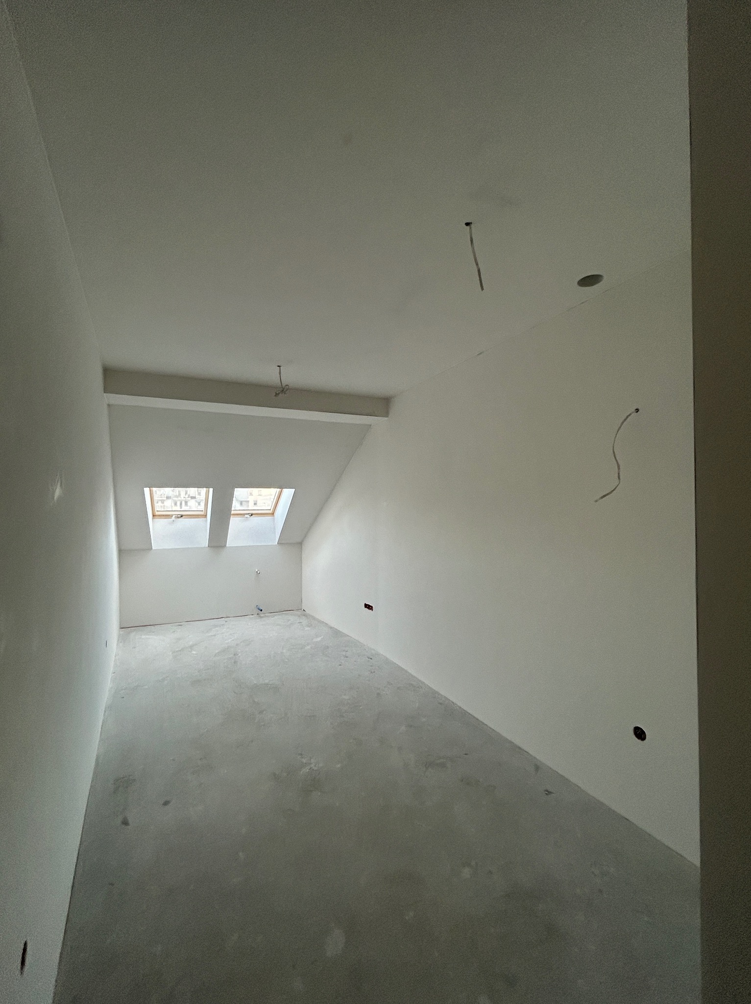 Wnętrze poddasza po gładziowaniu ścian i sufitu, widoczne dwa okna dachowe i wystające przewody elektryczne. Szare wykończenie podłogi.