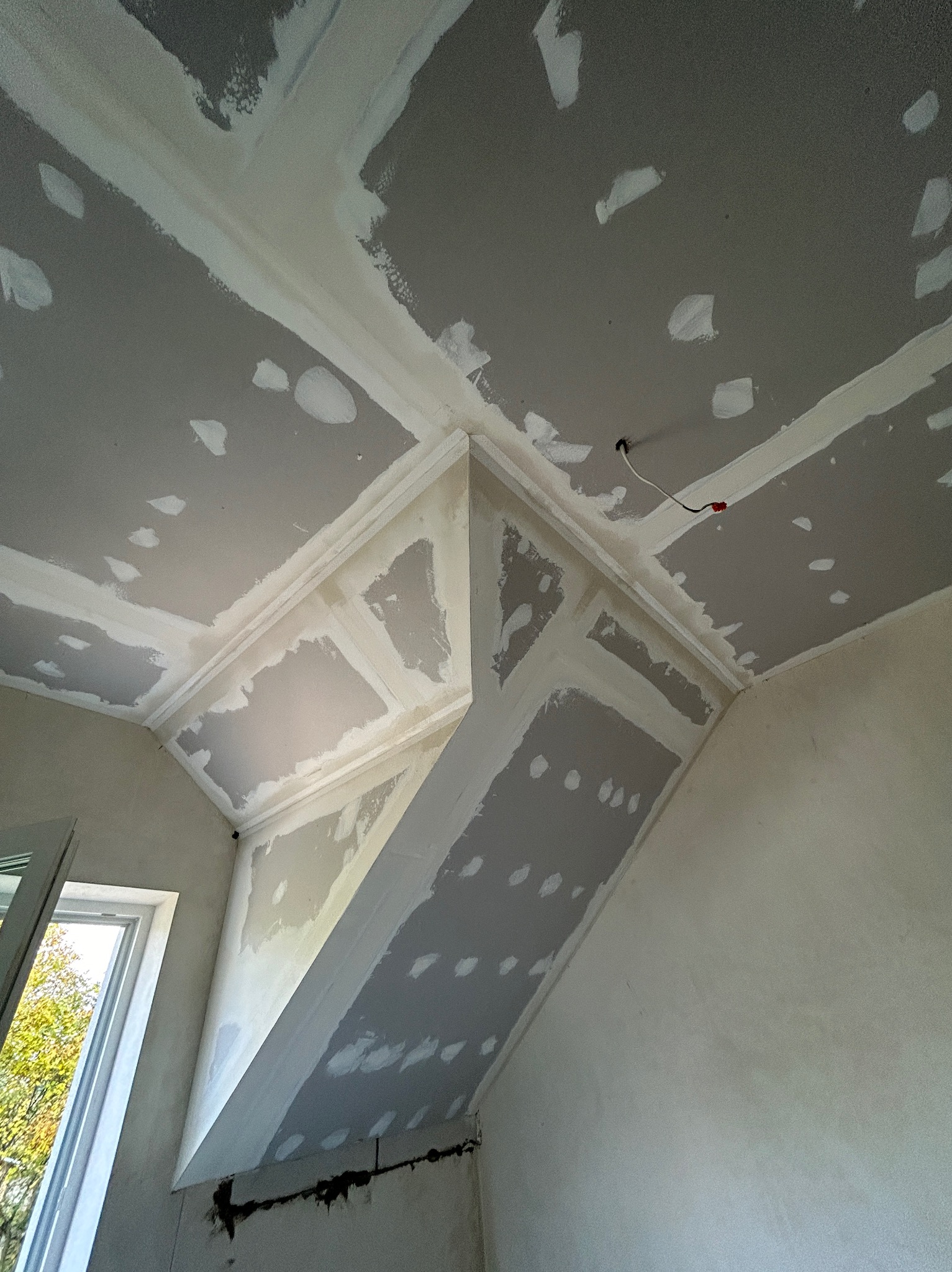 Skośny sufit poddasza po montażu płyt gipsowo-kartonowych, z widocznymi łączeniami i szpachlowaniem. Widok z dołu na fragment okna i drzewo.