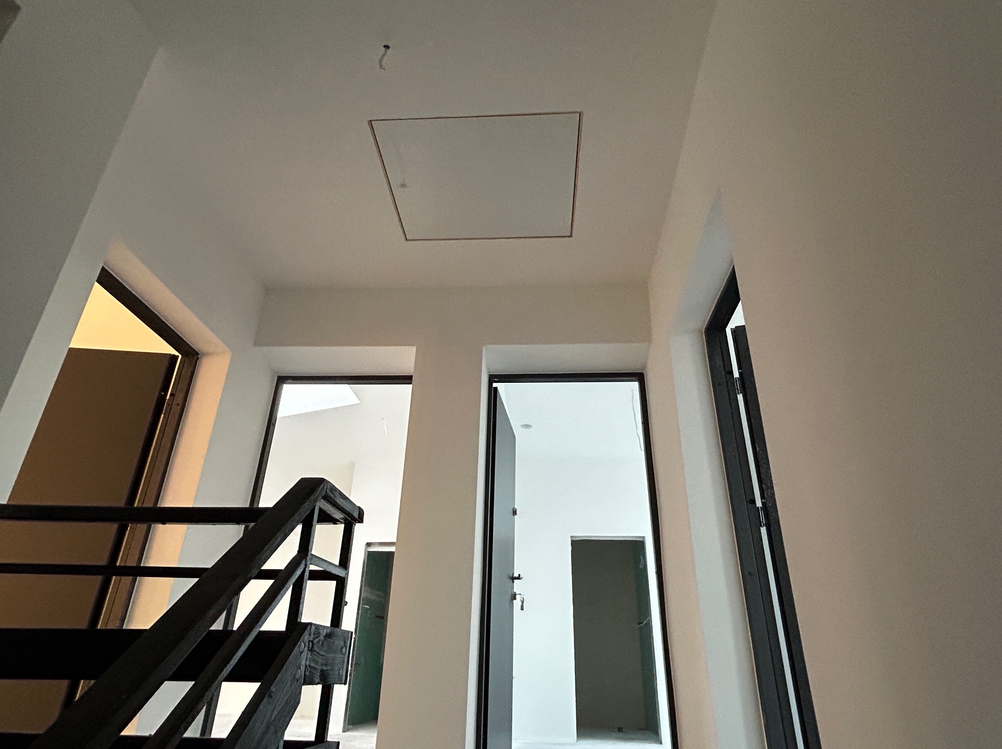 Wnętrze w trakcie remontu: białe ściany, otwarte drzwi z czarnymi futrynami, drewniane schody z czarną balustradą, klapa rewizyjna na suficie. Widok z dołu.