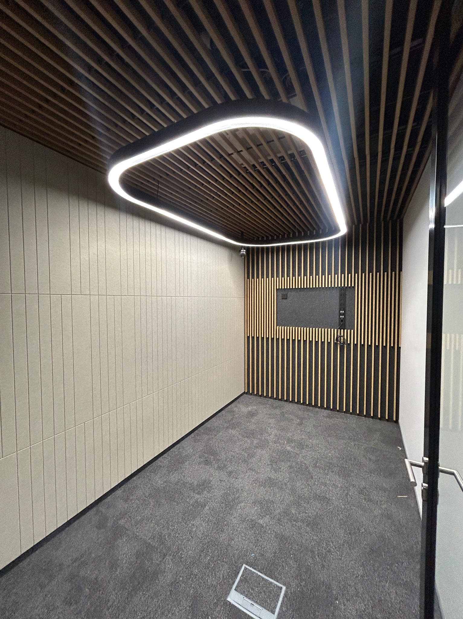 Nowoczesne wykończenie biura: drewniany sufit z oświetleniem LED, panelowa ściana i ciemna wykładzina. Widoczny fragment monitora na ścianie z drewnianymi listwami.