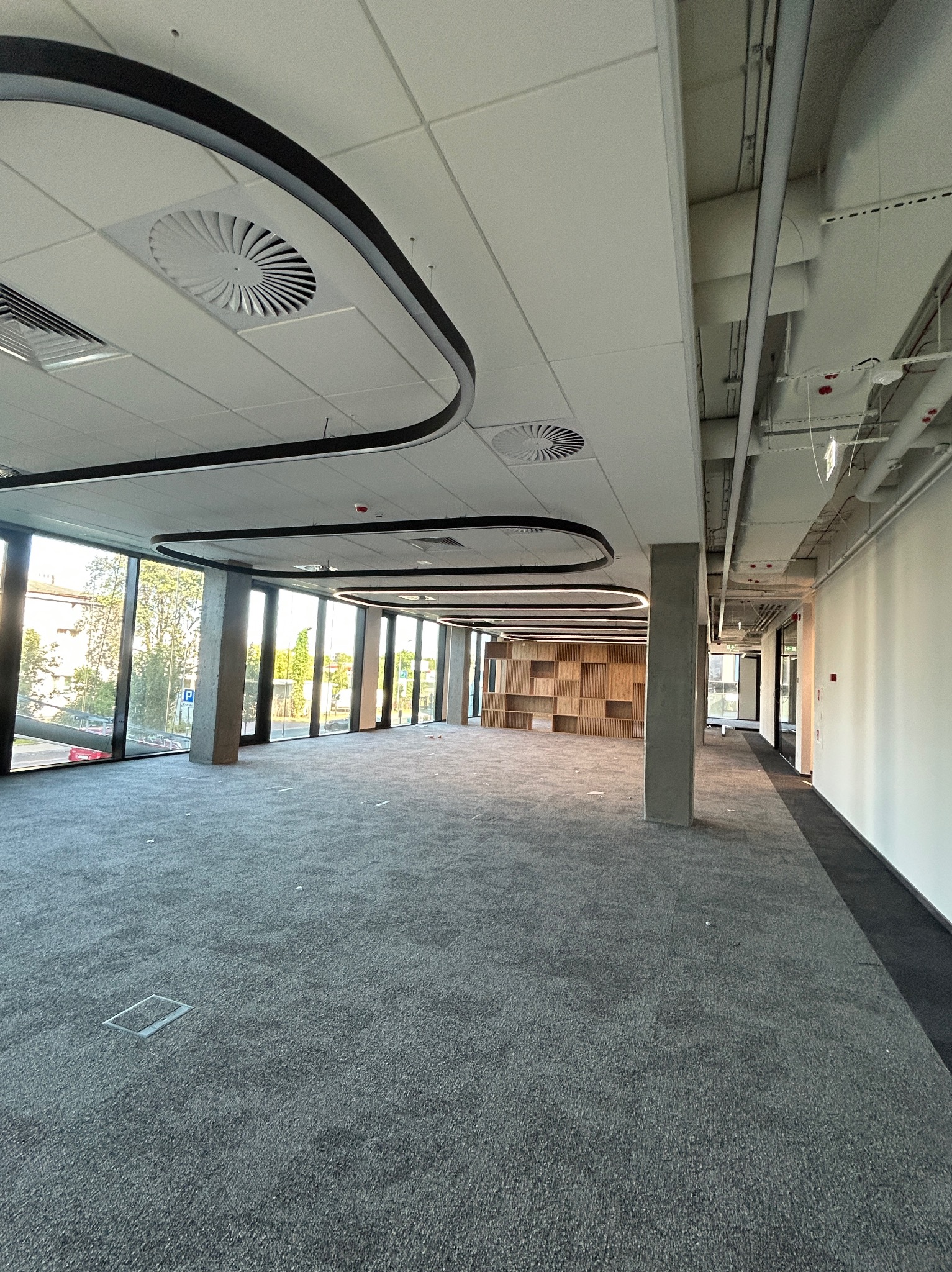 Przestronne wnętrze biurowe z szarą wykładziną, dużymi oknami i nowoczesnym oświetleniem sufitowym. Widoczne instalacje pod sufitem i elementy wykończeniowe.