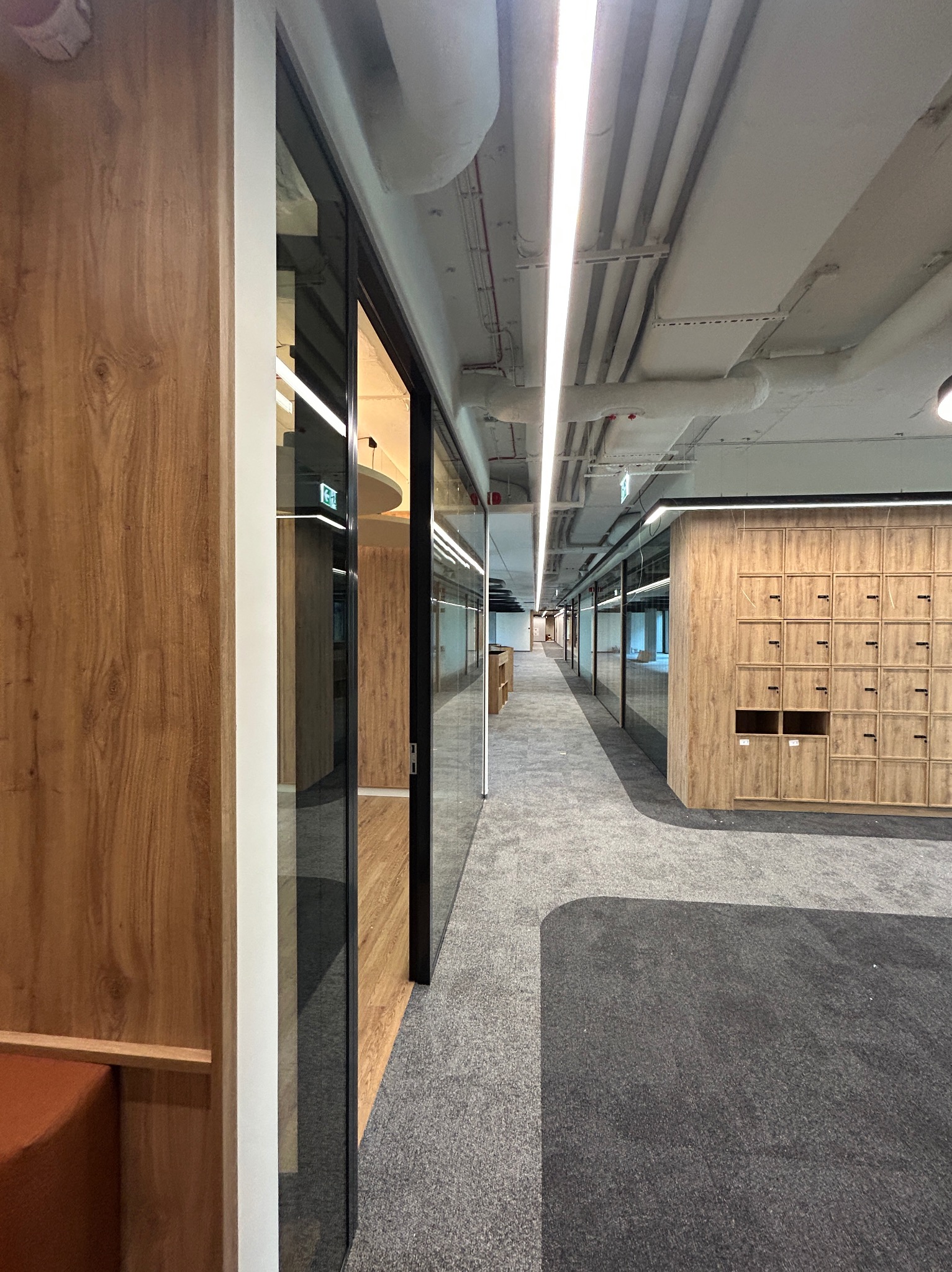 Nowoczesne wykończenie biura z drewnianymi akcentami, szarą wykładziną i przeszklonymi ścianami. Widoczne szafki pracownicze i oświetlenie sufitowe.