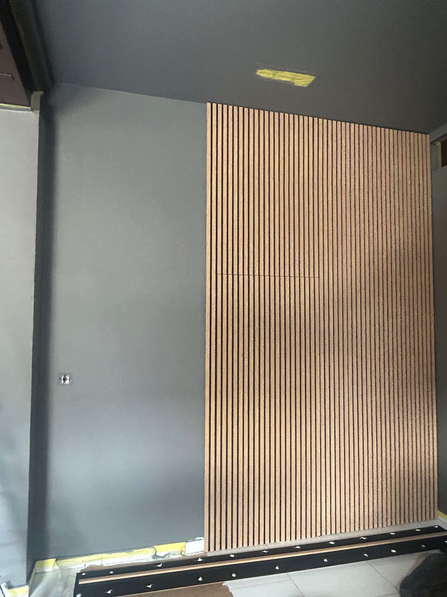 Szara ściana z drewnianymi panelami akustycznymi, widoczne łączenia paneli. Fragment podłogi z zabezpieczeniami. Ciemnoszary sufit z wbudowanym oświetleniem.