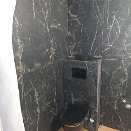 Robert Ziółkowski Usługi Ogólnobudowlane - Wykończona łazienka z czarną toaletą i marmurowymi płytkami na ścianach, widok na podłogę z paneli imitujących drewno.