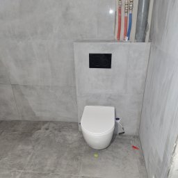 Robert Ziółkowski Usługi Ogólnobudowlane - Montaż podwieszanej toalety z szarą płytką imitującą beton, widoczne rury instalacji wodnej i kanalizacyjnej, oraz czarny przycisk spłuczki.