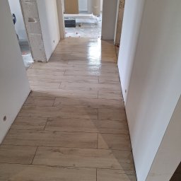 Robert Ziółkowski Usługi Ogólnobudowlane - Korytarz w trakcie prac wykończeniowych, świeżo ułożona podłoga z paneli imitujących drewno, widoczne białe ściany w stanie surowym, w tle otwarte przejście do innego pomieszczenia z widocznymi...