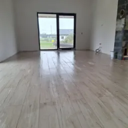 Jasne panele podłogowe imitujące drewno, ułożone w pustym, surowym pomieszczeniu z dużym oknem balkonowym i fragmentem obudowy kominka w trakcie prac wykończeniowych.