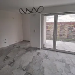 Wnętrze pokoju z marmurową posadzką, nowoczesną lampą i dużym oknem balkonowym. Widok na blok mieszkalny z balkonu. Świeżo wykończone, jasne pomieszczenie.