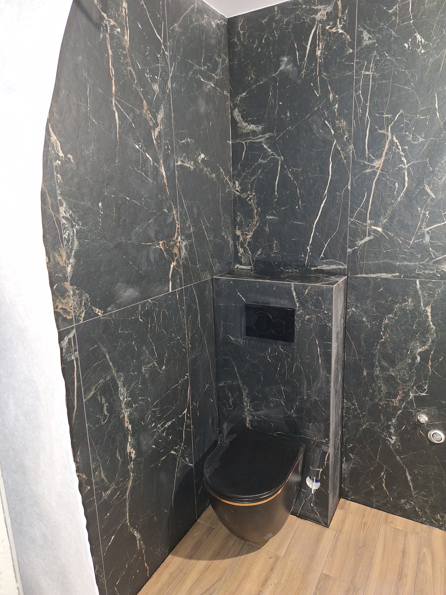 Wykończona łazienka z czarną toaletą i marmurowymi płytkami na ścianach, widok na podłogę z paneli imitujących drewno.