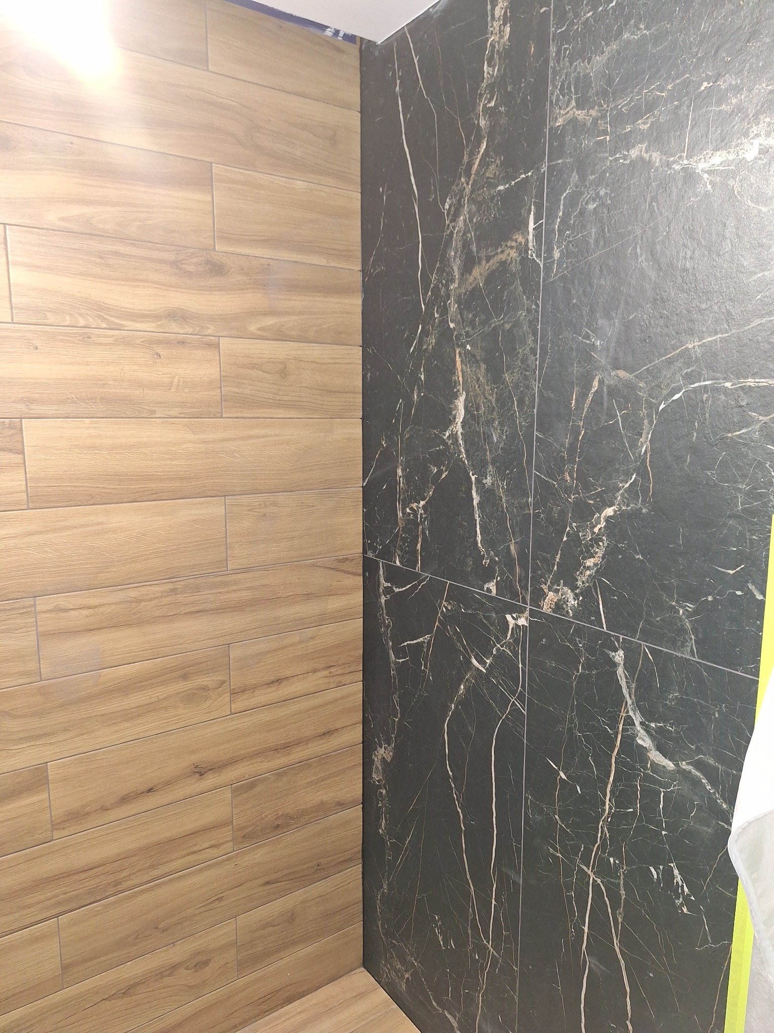 Ściana wyłożona dwoma rodzajami płytek: imitacją drewna w odcieniach brązu oraz czarnymi płytkami z białym marmurkowym wzorem, widoczne łączenia między płytkami.