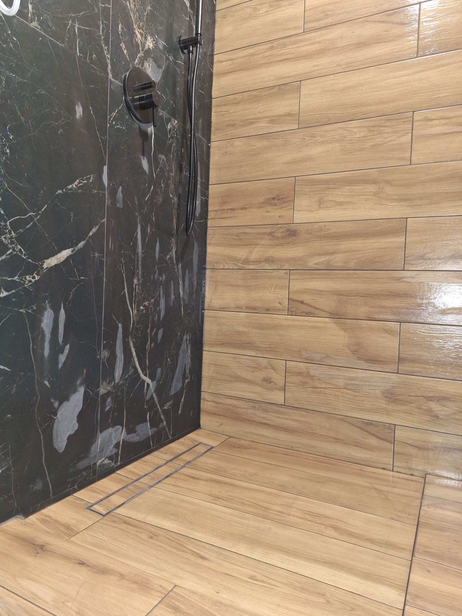 Nowoczesna kabina prysznicowa z czarną marmurową ścianą i drewnopodobnymi płytkami, z odpływem liniowym.