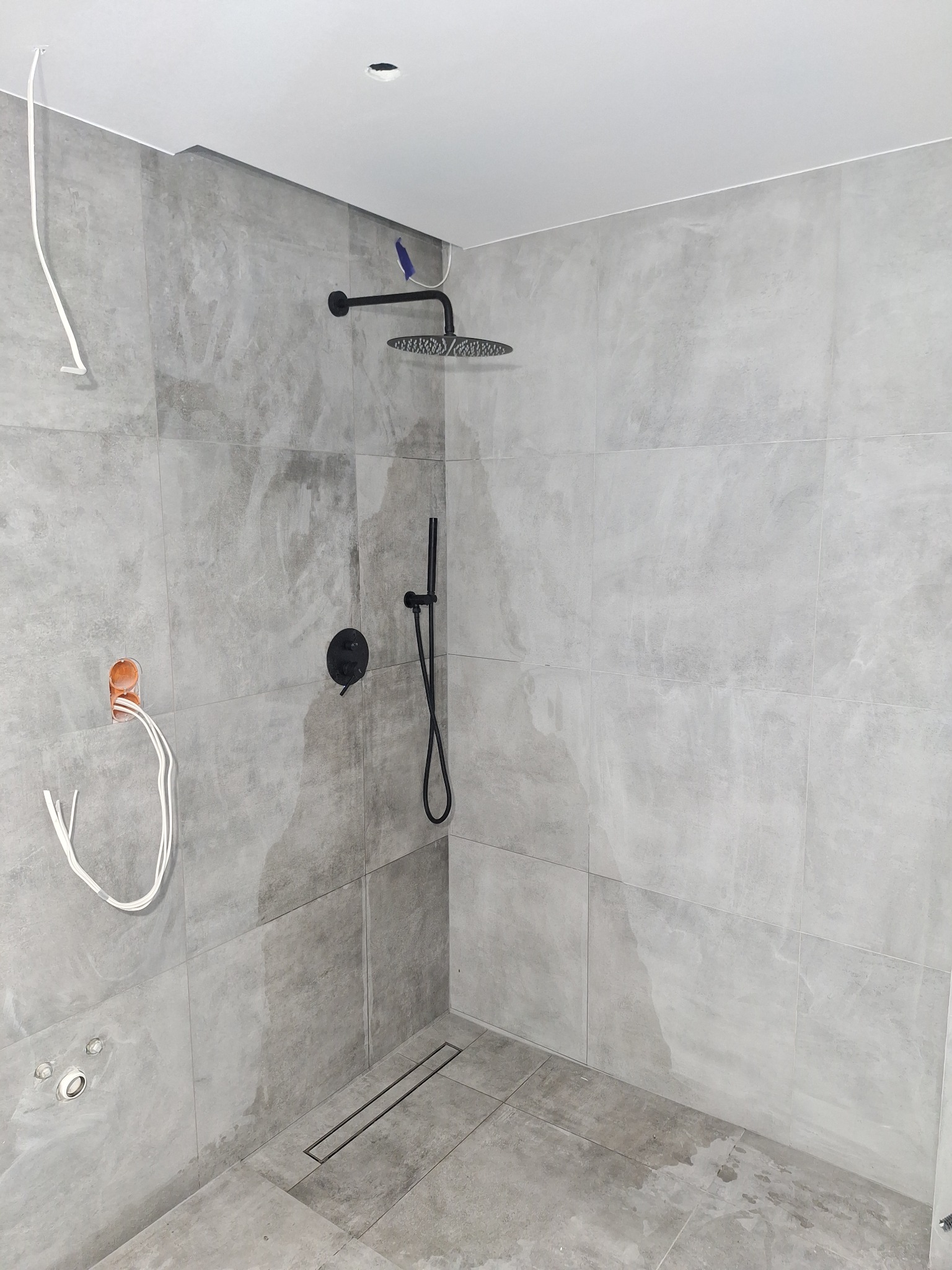 Nowoczesny prysznic typu walk-in z szarą, betonową okładziną ścienną, czarną armaturą i odpływem liniowym, w trakcie montażu widoczne wystające przewody elektryczne.