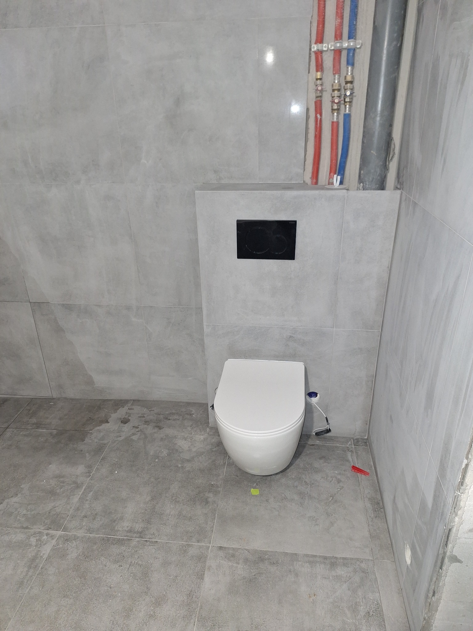 Montaż podwieszanej toalety z szarą płytką imitującą beton, widoczne rury instalacji wodnej i kanalizacyjnej, oraz czarny przycisk spłuczki.