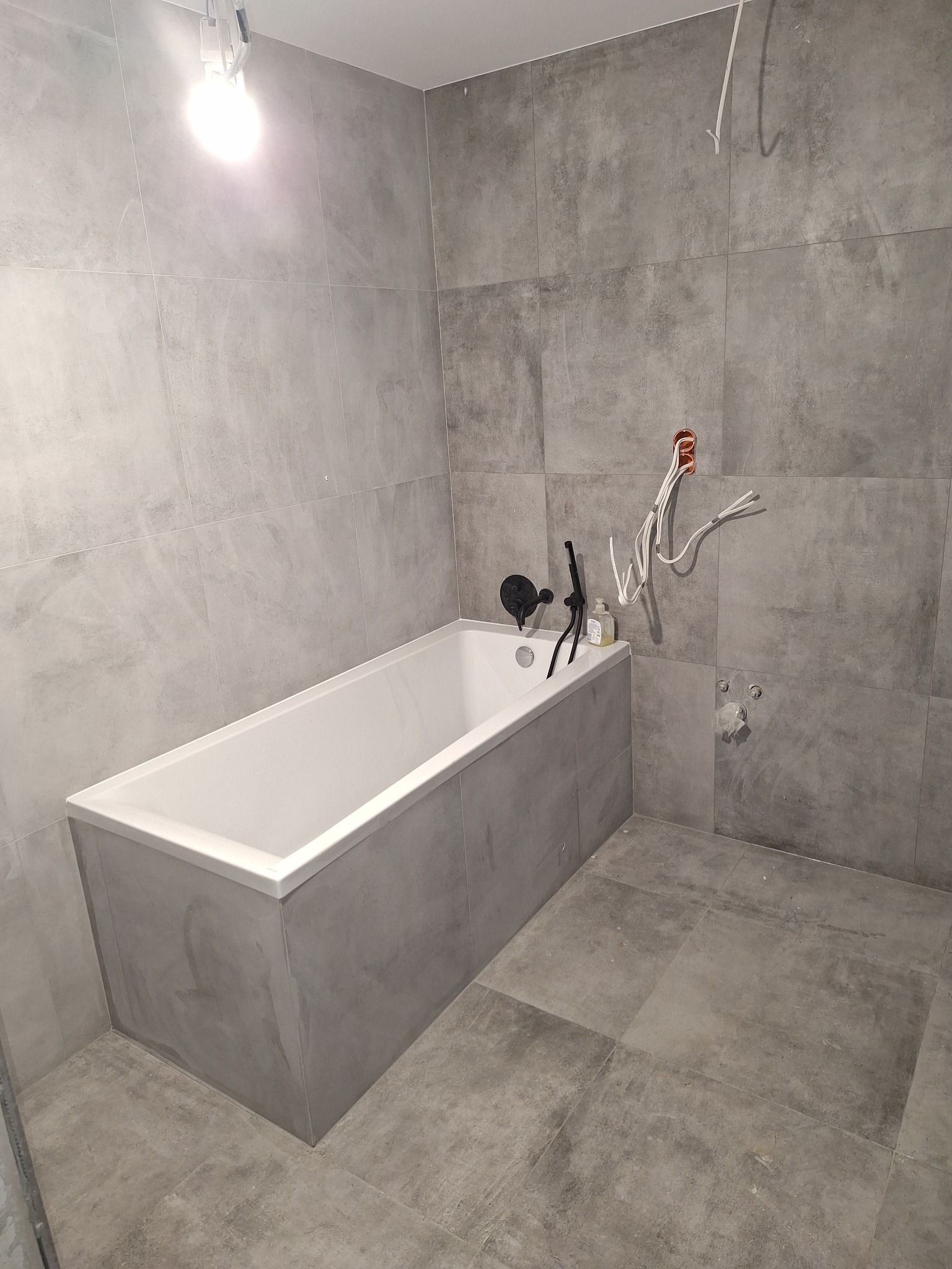 Łazienka w trakcie wykańczania: biała wanna obudowana szarymi płytkami, szare płytki na ścianach i podłodze, widoczne przewody elektryczne i instalacja wodna przed podłączeniem armatury.