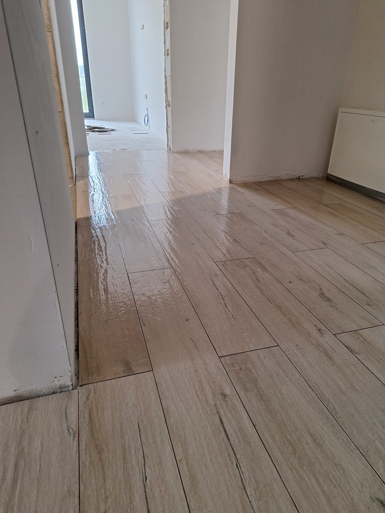 Świeżo ułożone panele podłogowe imitujące drewno, mokre od mycia, w jasnym, surowym wnętrzu z widocznymi niedokończonymi ścianami i fragmentami podłoża w tle.