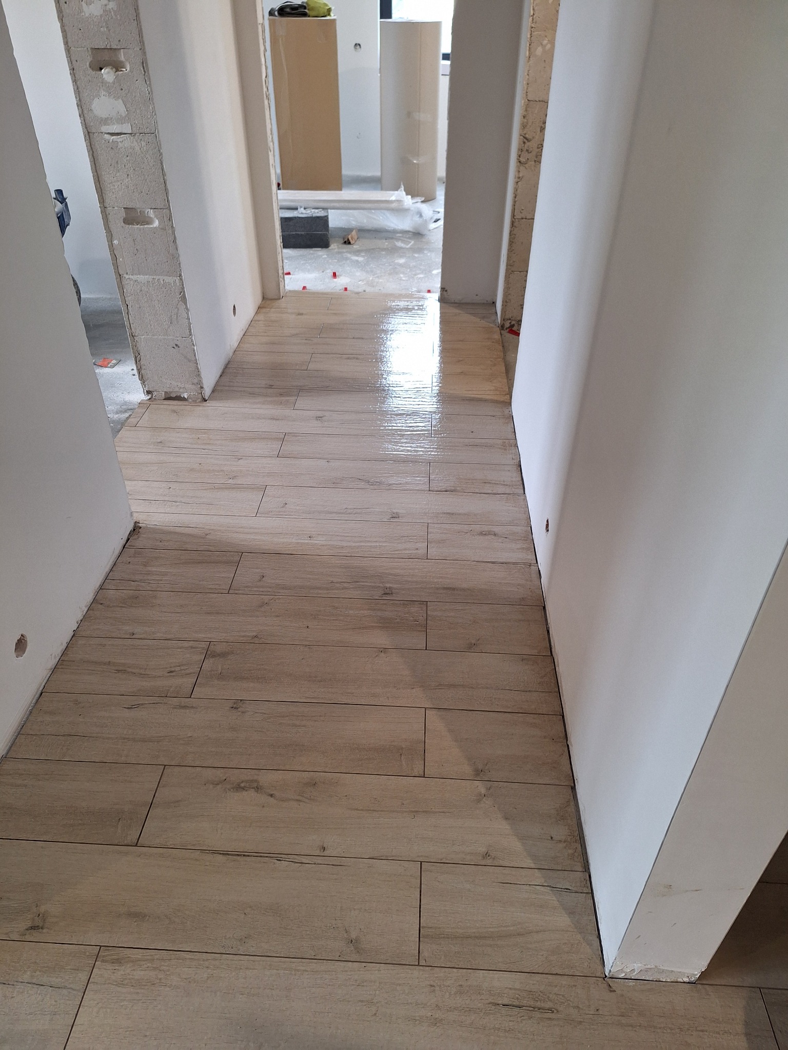 Korytarz w trakcie prac wykończeniowych, świeżo ułożona podłoga z paneli imitujących drewno, widoczne białe ściany w stanie surowym, w tle otwarte przejście do innego pomieszczenia z widocznymi...