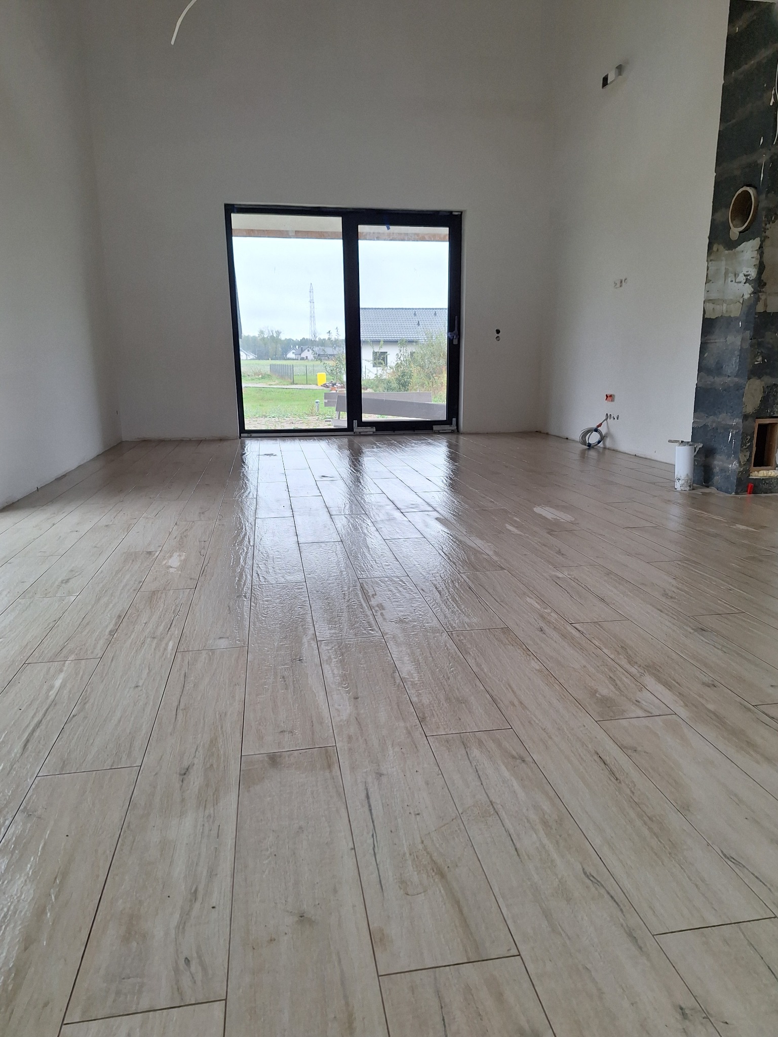 Jasne panele podłogowe imitujące drewno, ułożone w pustym, surowym pomieszczeniu z dużym oknem balkonowym i fragmentem obudowy kominka w trakcie prac wykończeniowych.