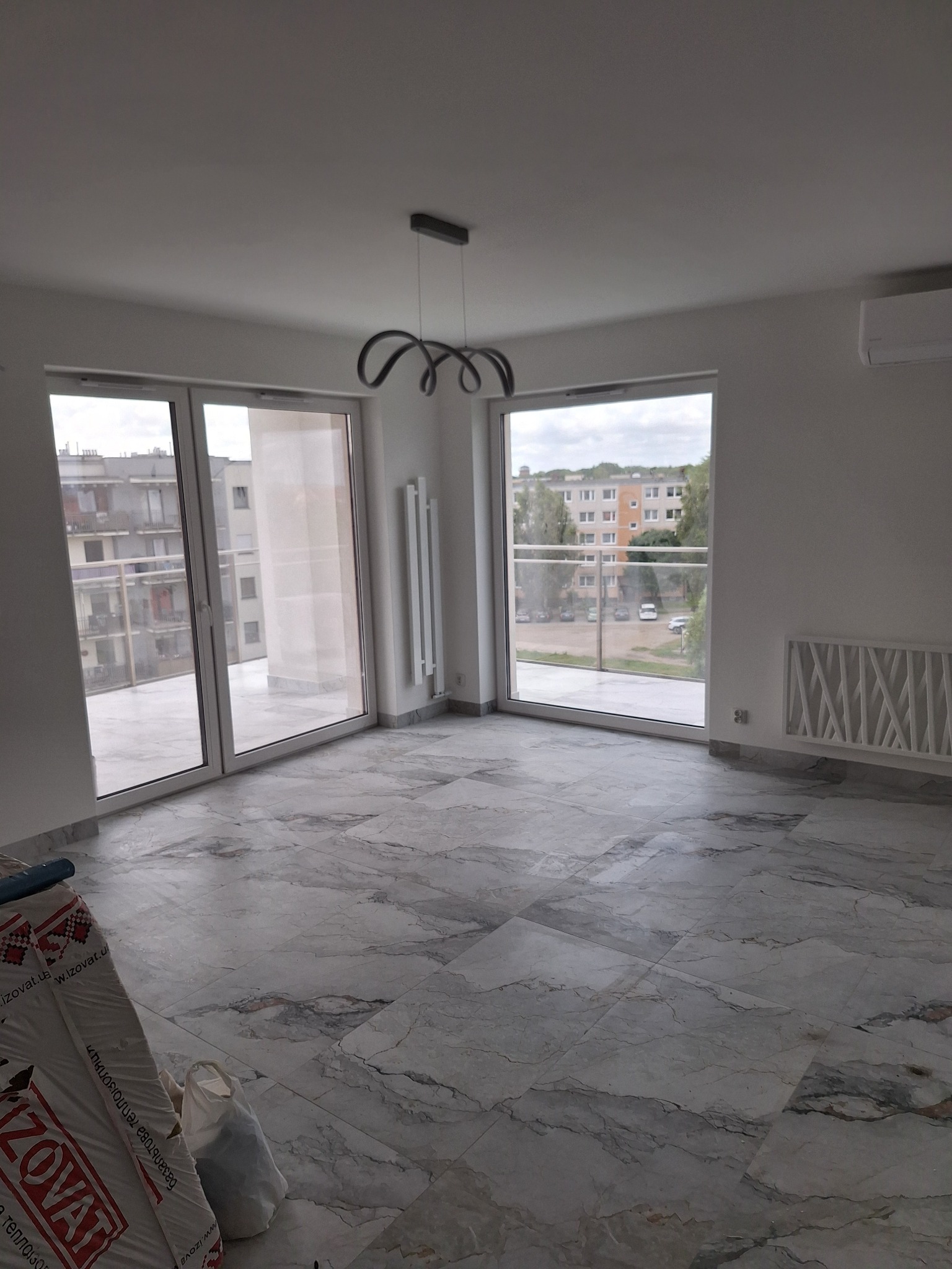 Jasne wnętrze po remoncie: marmurowe płytki podłogowe, duże okna balkonowe z widokiem na osiedle, nowoczesna lampa sufitowa i grzejnik dekoracyjny.