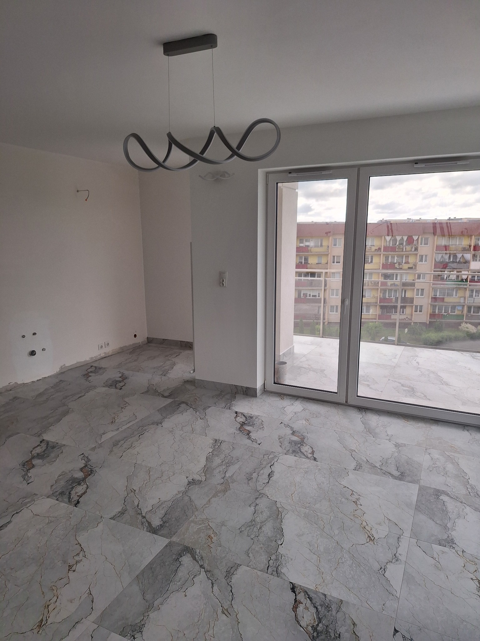 Wnętrze pokoju z marmurową posadzką, nowoczesną lampą i dużym oknem balkonowym. Widok na blok mieszkalny z balkonu. Świeżo wykończone, jasne pomieszczenie.