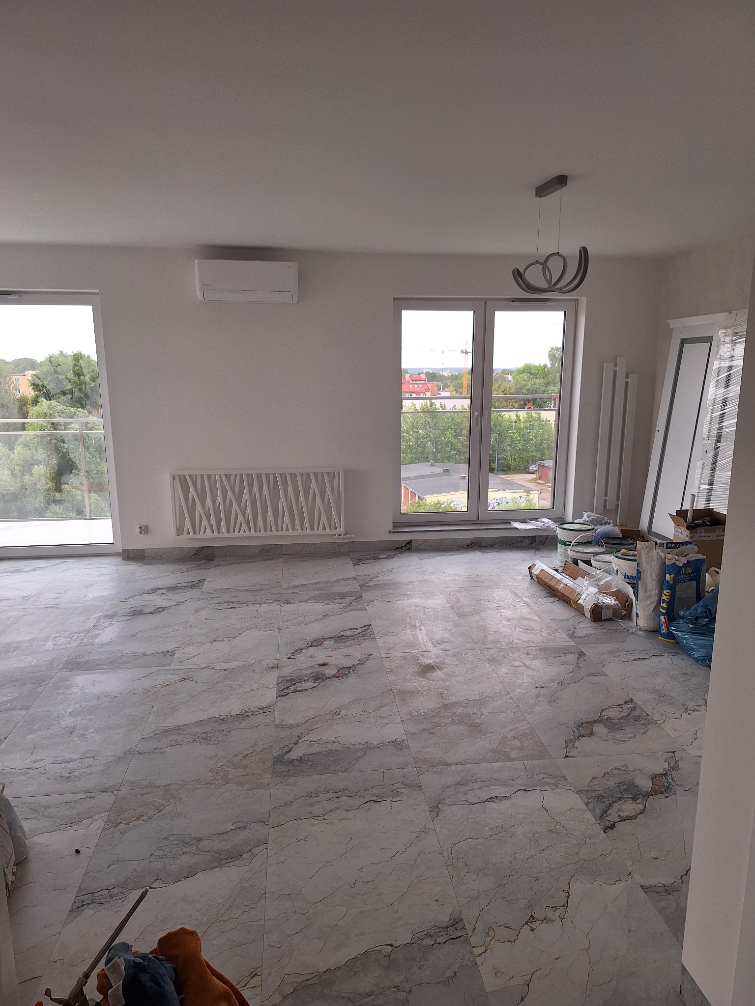 Jasne wnętrze z marmurową podłogą, grzejnikiem dekoracyjnym i nowoczesną lampą. Widoczne okna z widokiem na zieleń i elementy wykończeniowe.