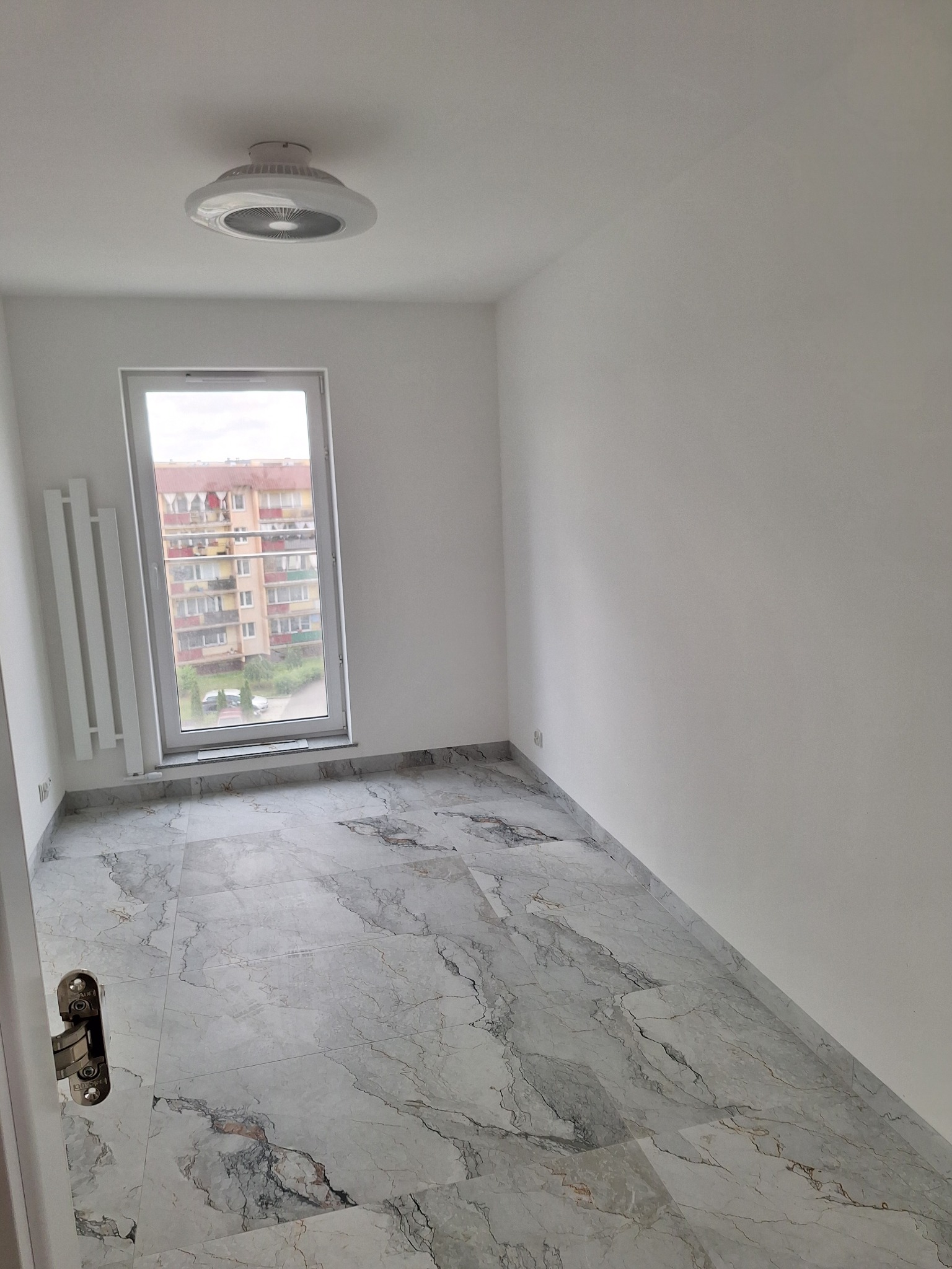 Jasne wnętrze po remoncie: białe ściany, marmurowe płytki podłogowe, okno z widokiem na budynek mieszkalny, nowoczesny grzejnik i designerska lampa sufitowa.