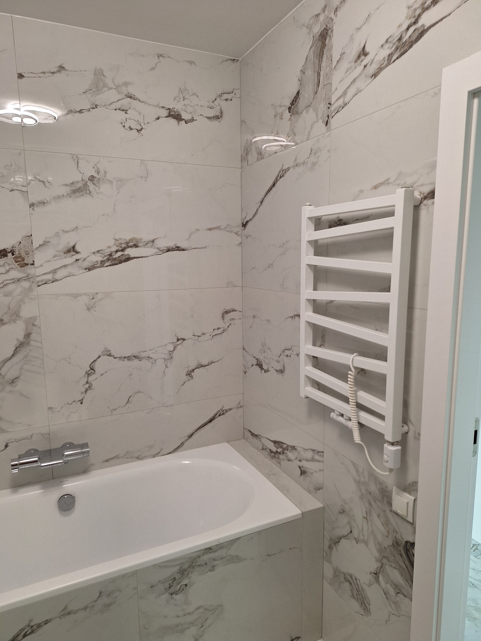 Biała wanna z baterią i grzejnik drabinkowy na ścianie wyłożonej marmurowymi płytkami w łazience. Nowoczesne wykończenie, minimalistyczny styl.