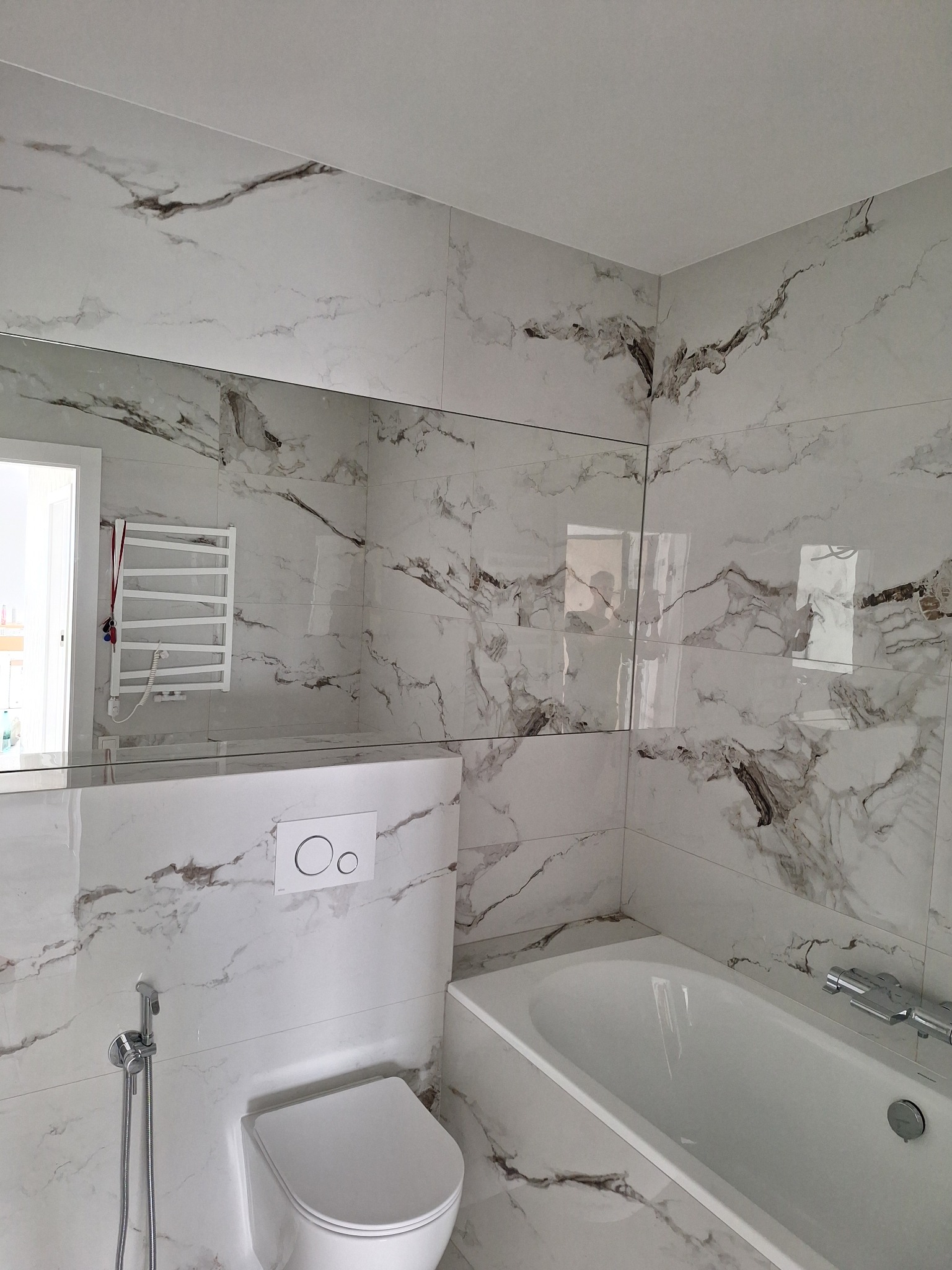 Wykończona łazienka z płytkami imitującymi marmur, biała wanna i toaleta. Widoczne detale: bateria wannowa, bidetka i grzejnik drabinkowy odbijający się w lustrze.