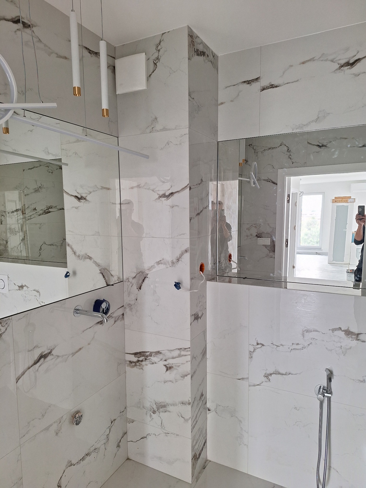 Wykończona łazienka z białymi płytkami imitującymi marmur, lustrami na ścianach i nowoczesnym oświetleniem. Widoczna armatura i fragment okna w odbiciu.