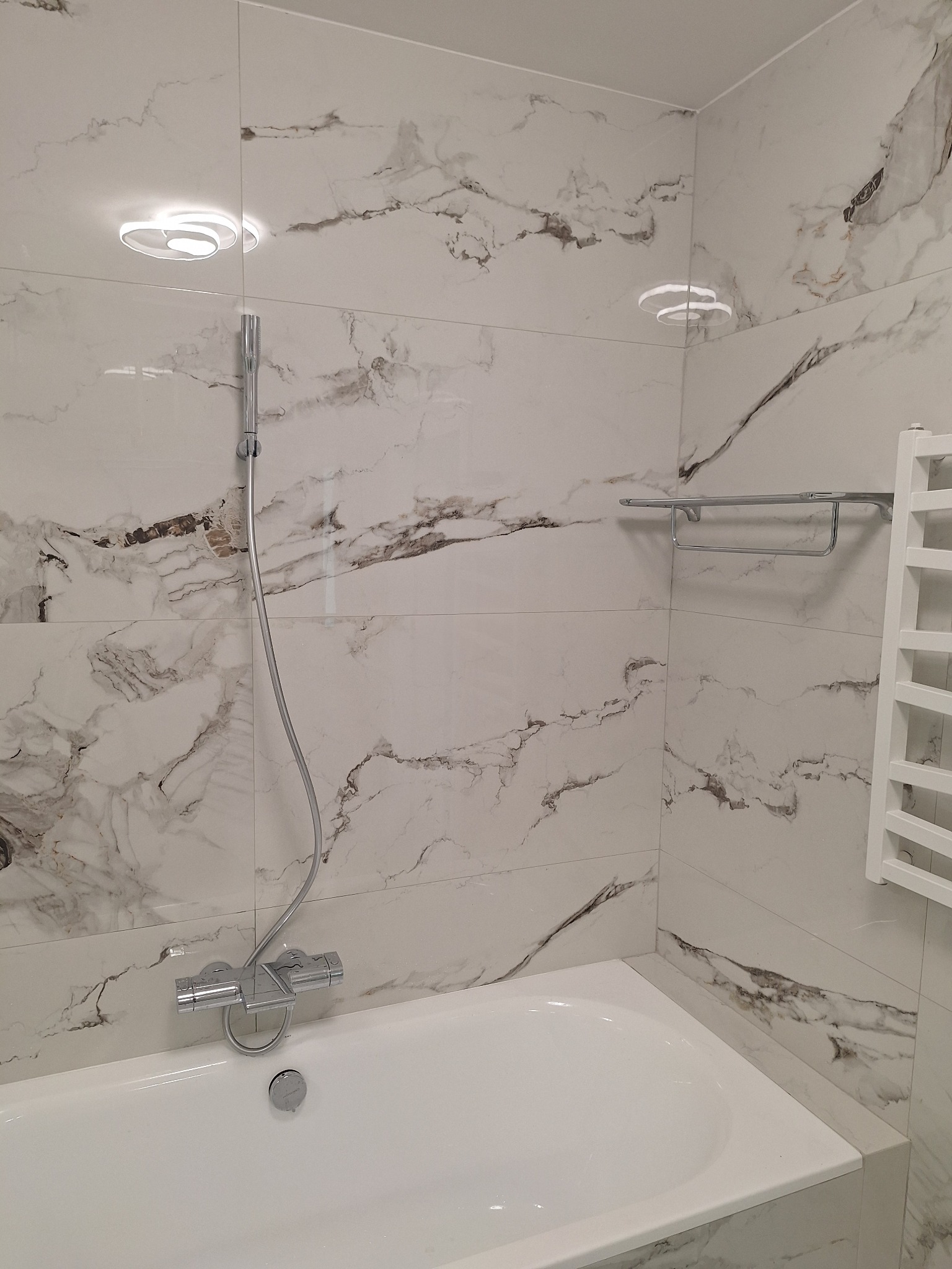 Wykończona łazienka z wanną, baterią natryskową i grzejnikiem drabinkowym na ścianie z płytek imitujących marmur. Nowoczesne oświetlenie sufitowe.