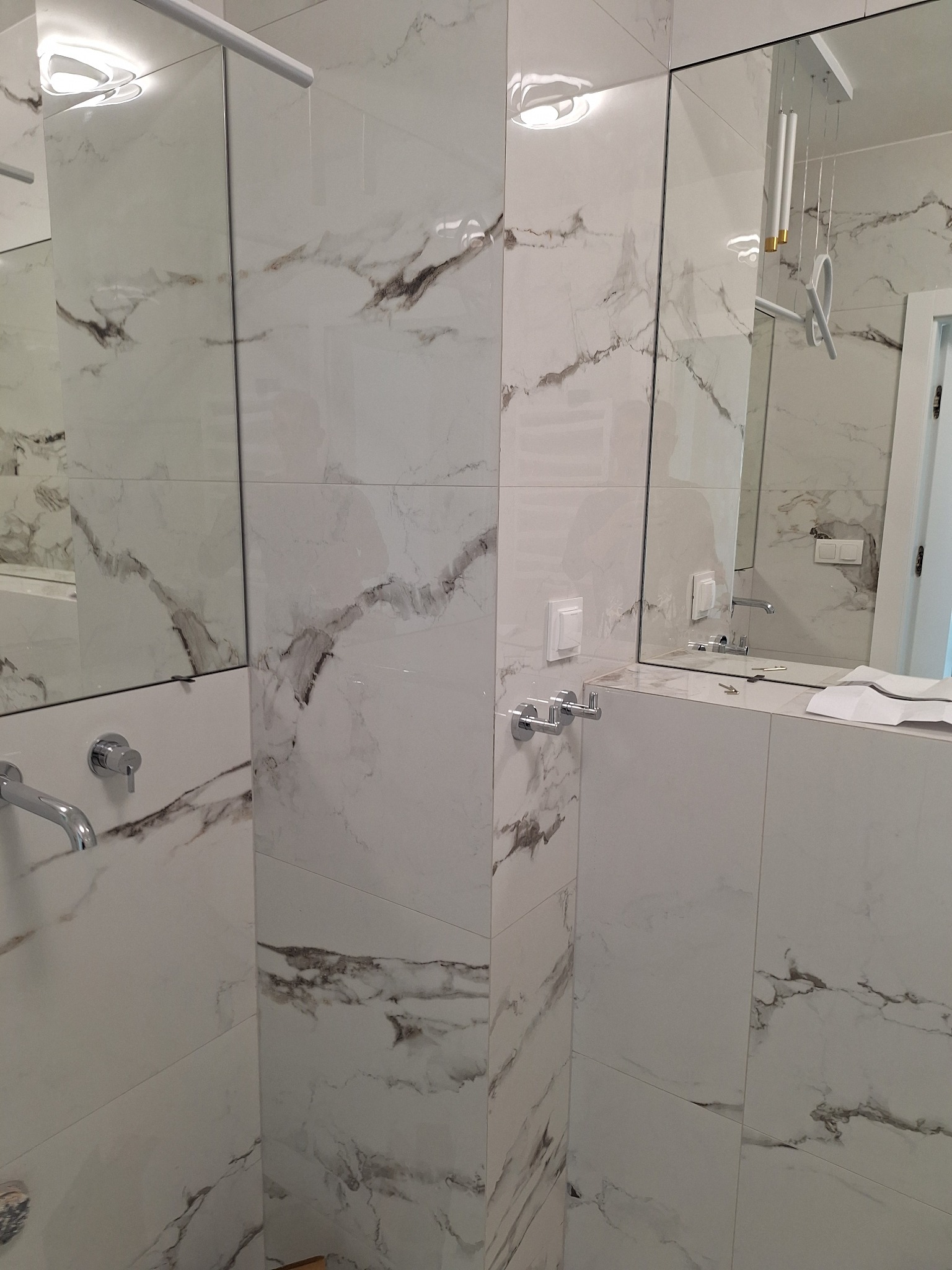 Biała łazienka z płytkami imitującymi marmur, chromowana armatura, duże lustra, nowoczesne oświetlenie. Widok na umywalkę i fragment ściany.