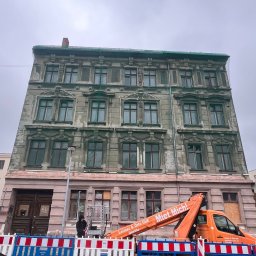 Sergiusz Dekarz - Renowacja elewacji kamienicy z podnośnika koszowego, zabezpieczona barierkami. Widoczny fragment samochodu. Pochmurne niebo.