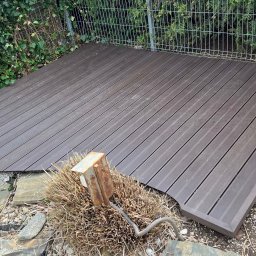 Sergiusz Dekarz - Ukończony taras z desek kompozytowych w ogrodzie, otoczony zielenią i kamieniami. Widoczny metalowy słupek i przewód. Deski ułożone równolegle, tworząc estetyczną przestrzeń.