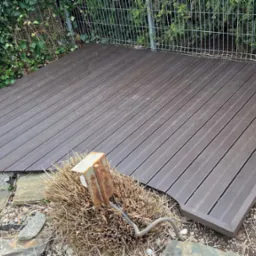 Ukończony taras z desek kompozytowych w ogrodzie, otoczony zielenią i kamieniami. Widoczny metalowy słupek i przewód. Deski ułożone równolegle, tworząc estetyczną przestrzeń.