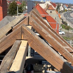 Sergiusz Dekarz - Konstrukcja więźby dachowej z drewnianymi belkami, widok z góry na tle zabudowy miejskiej w słoneczny dzień. Częściowo widoczna osoba w trakcie pracy.