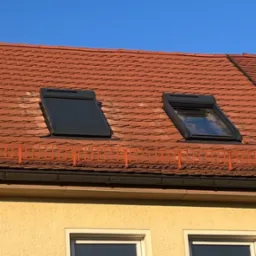 Czerwona dachówka z dwoma oknami dachowymi, jedno z roletą zewnętrzną. Fragment ściany budynku z dwoma oknami na dole kadru. Błękitne niebo w tle.