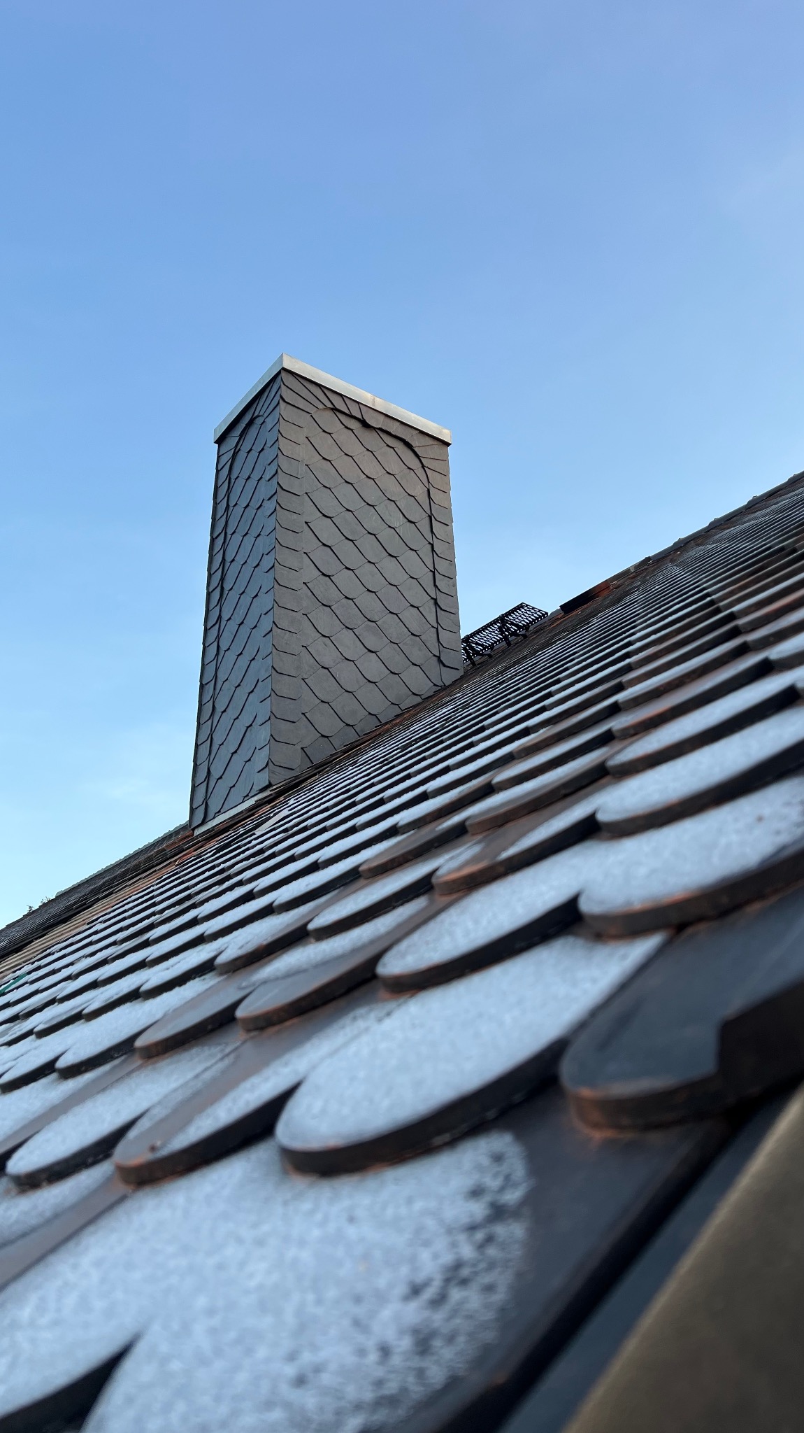 Dach pokryty okrągłymi dachówkami, lekko oszroniony, z kominem obłożonym łupkiem, na tle błękitnego nieba.