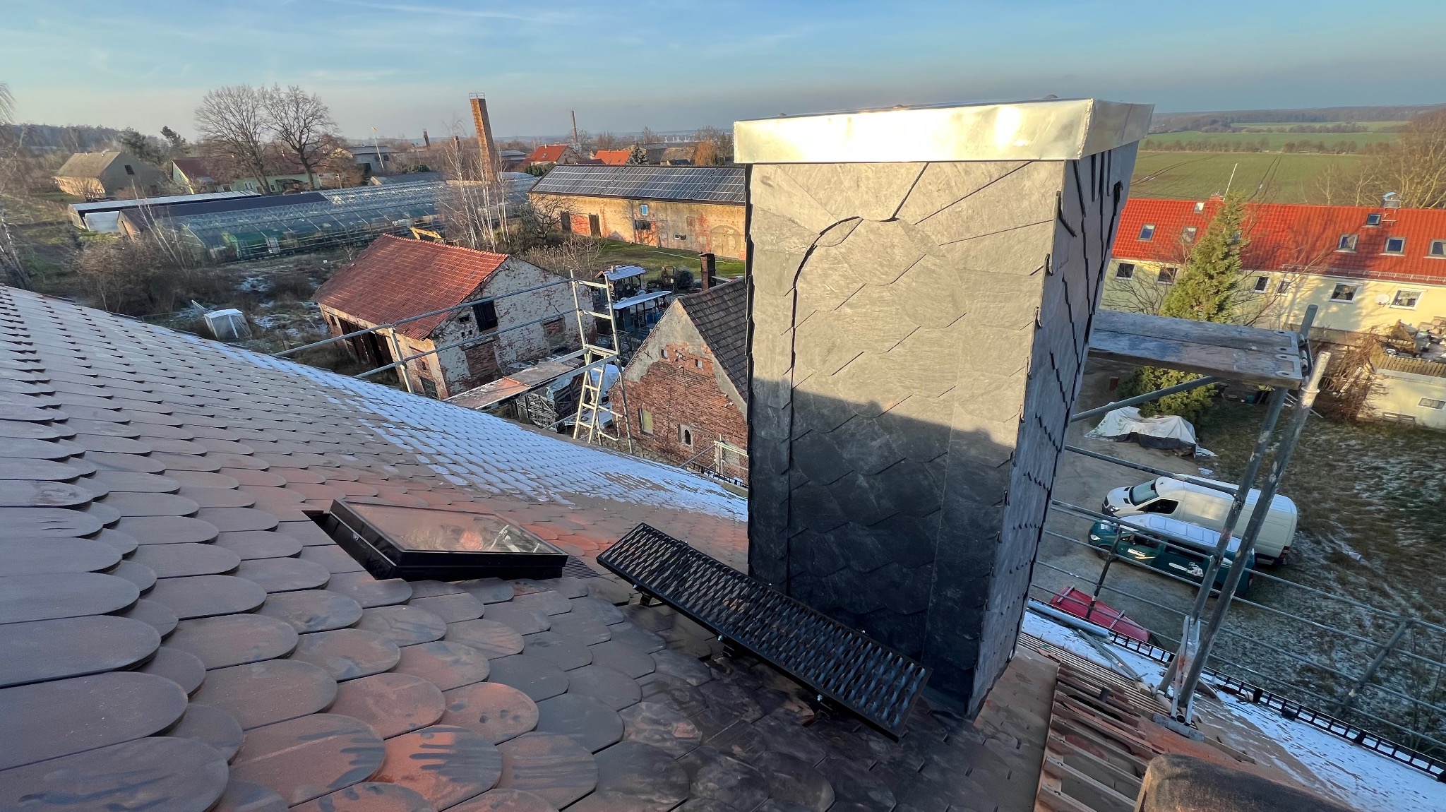 Widok z góry na dach z ceramiczną dachówką karpiówką, z zamontowanym oknem dachowym i metalową ławą kominiarską, komin obłożony łupkiem, w tle zabudowania gospodarcze i krajobraz wiejski.