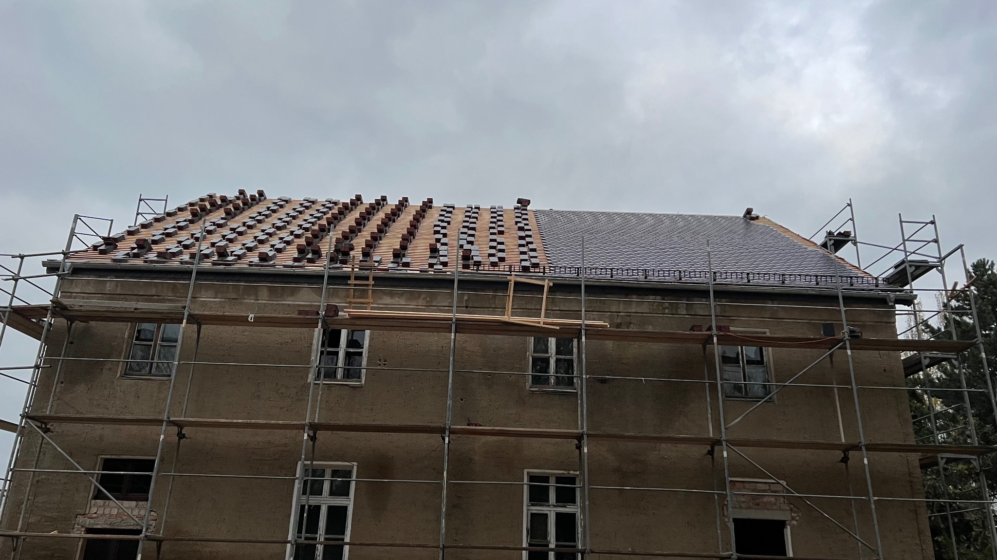 Budynek w trakcie układania dachówki, widoczne rusztowania i część dachu z już ułożoną dachówką oraz część z przygotowanymi dachówkami do ułożenia, pochmurne niebo w tle.