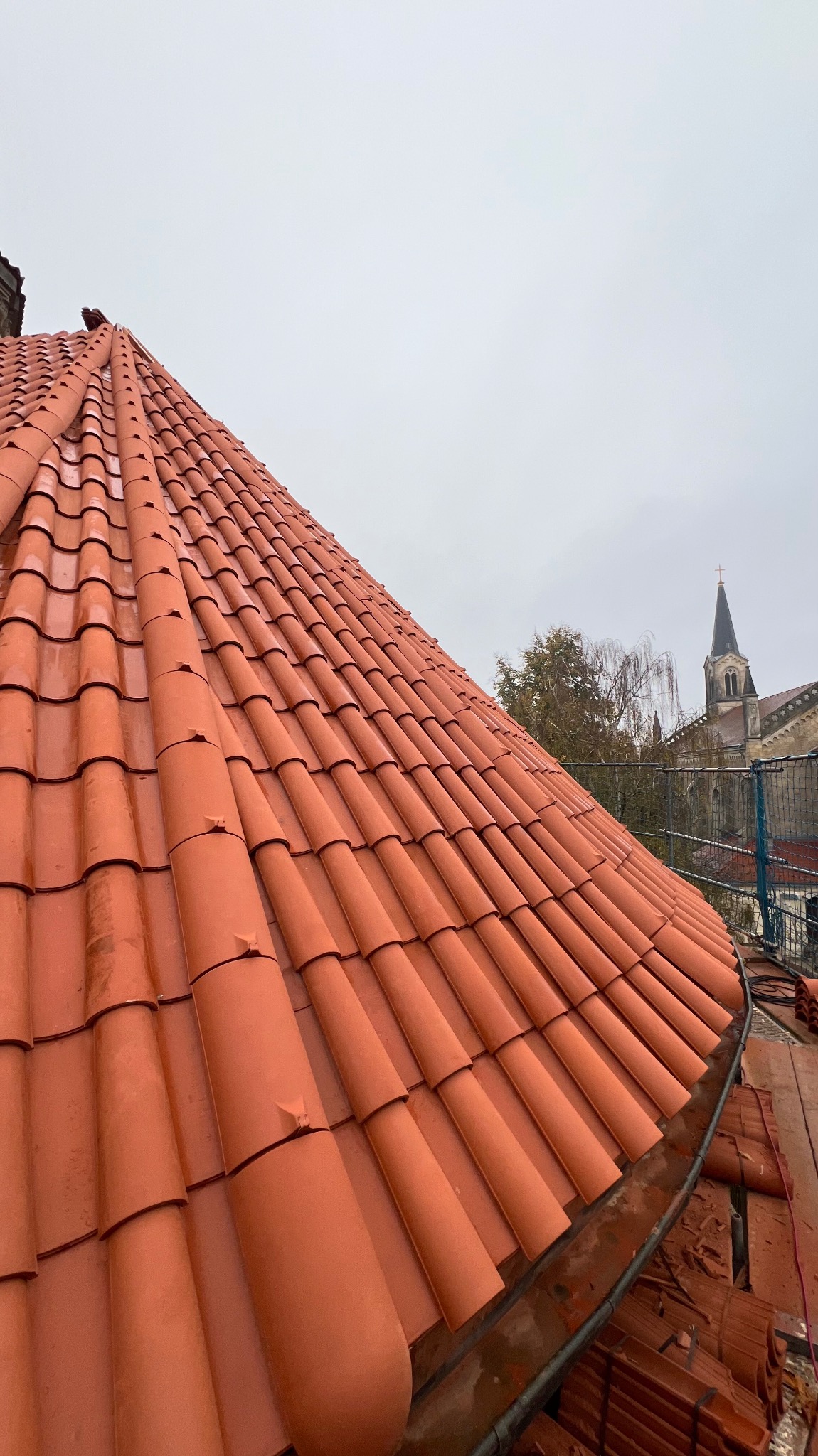 Czerwona dachówka ceramiczna na skośnym dachu, widok z góry, w tle zarys wieży kościelnej i pochmurne niebo.