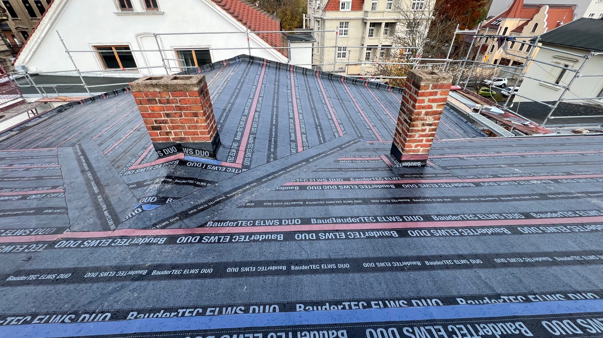 Montaż papy termozgrzewalnej BauderTEC ELWS DUO na dachu z dwoma kominami z cegły, widoczne rusztowania.