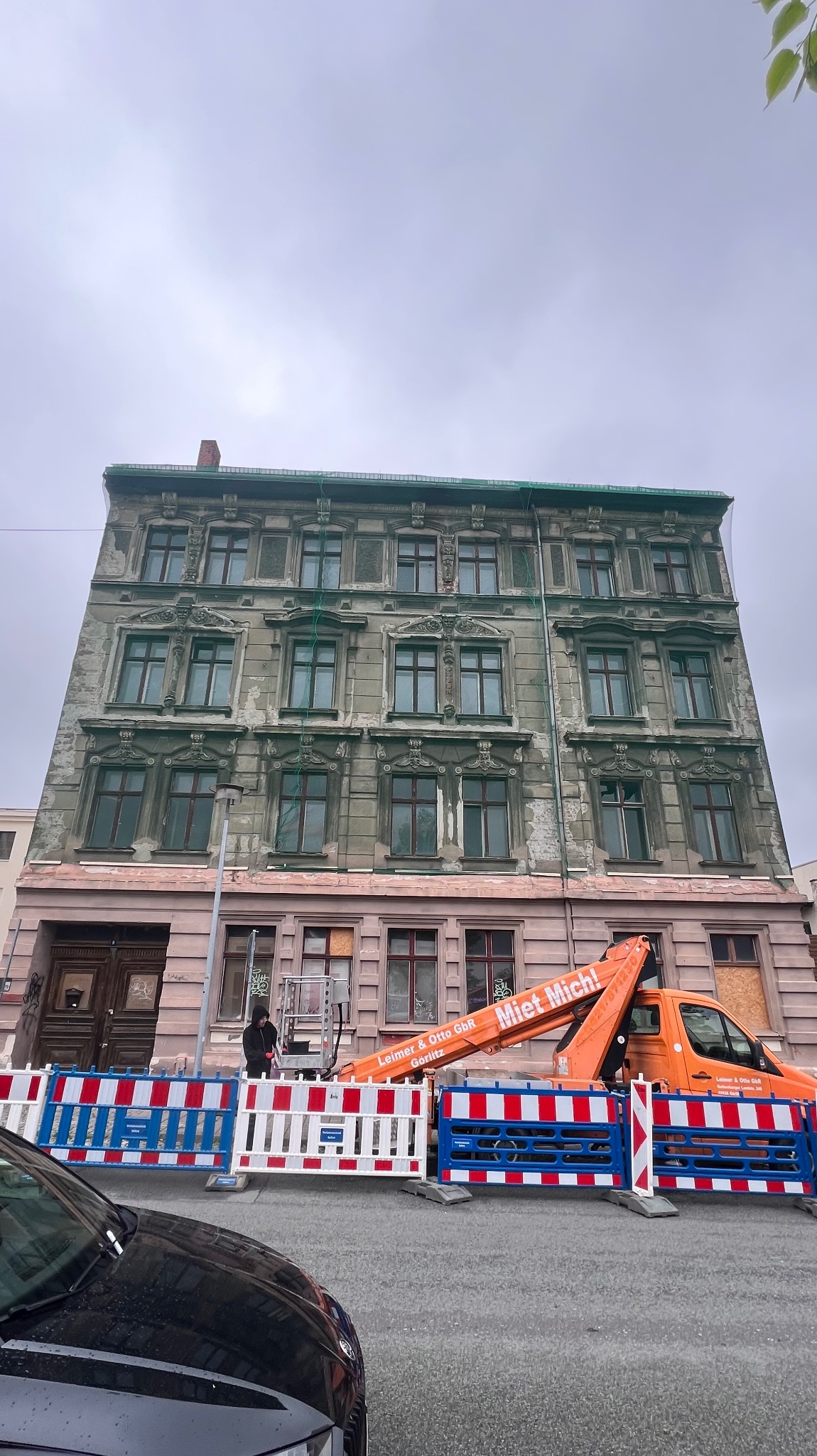Renowacja elewacji kamienicy z podnośnika koszowego, zabezpieczona barierkami. Widoczny fragment samochodu. Pochmurne niebo.