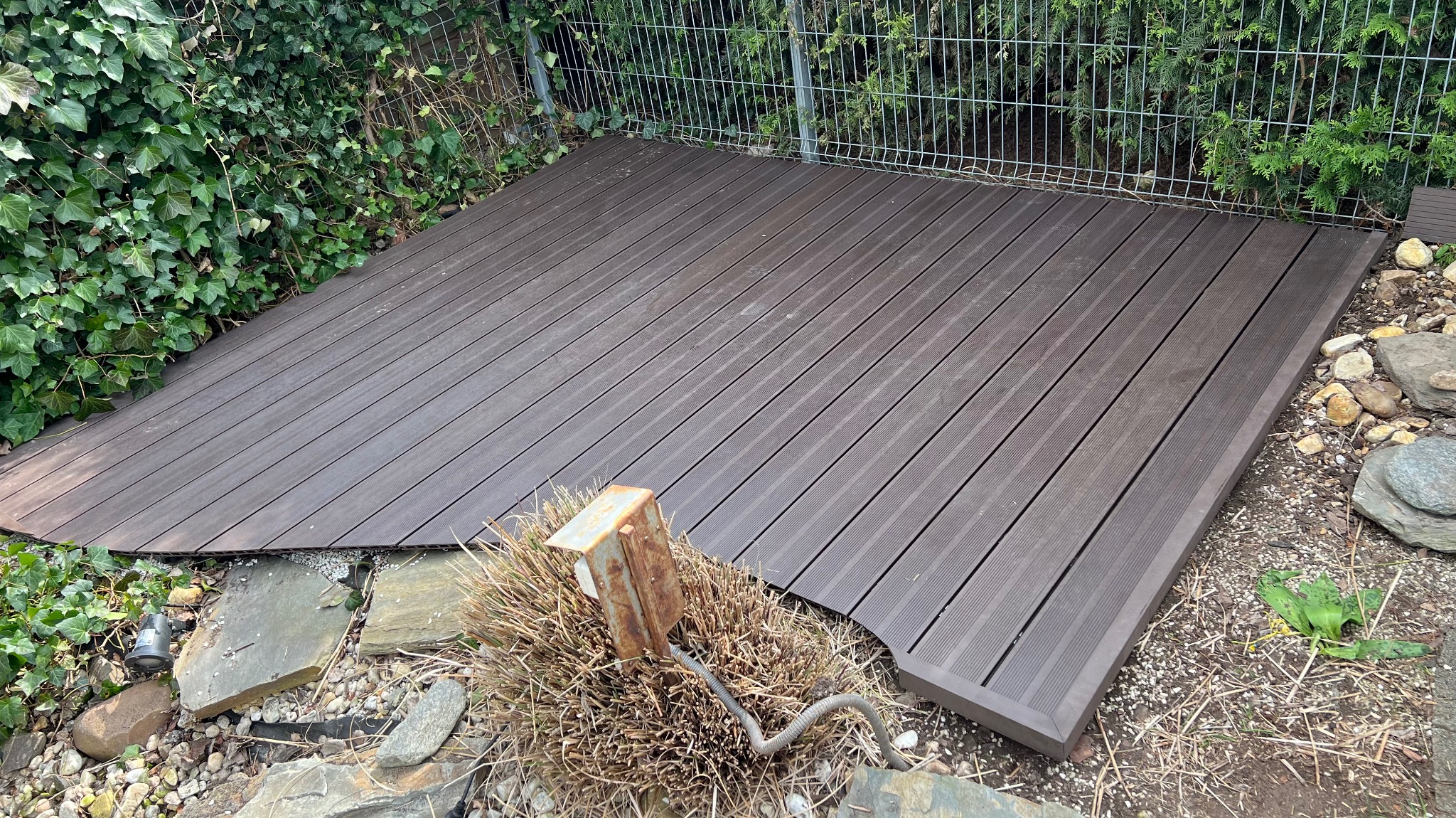 Ukończony taras z desek kompozytowych w ogrodzie, otoczony zielenią i kamieniami. Widoczny metalowy słupek i przewód. Deski ułożone równolegle, tworząc estetyczną przestrzeń.