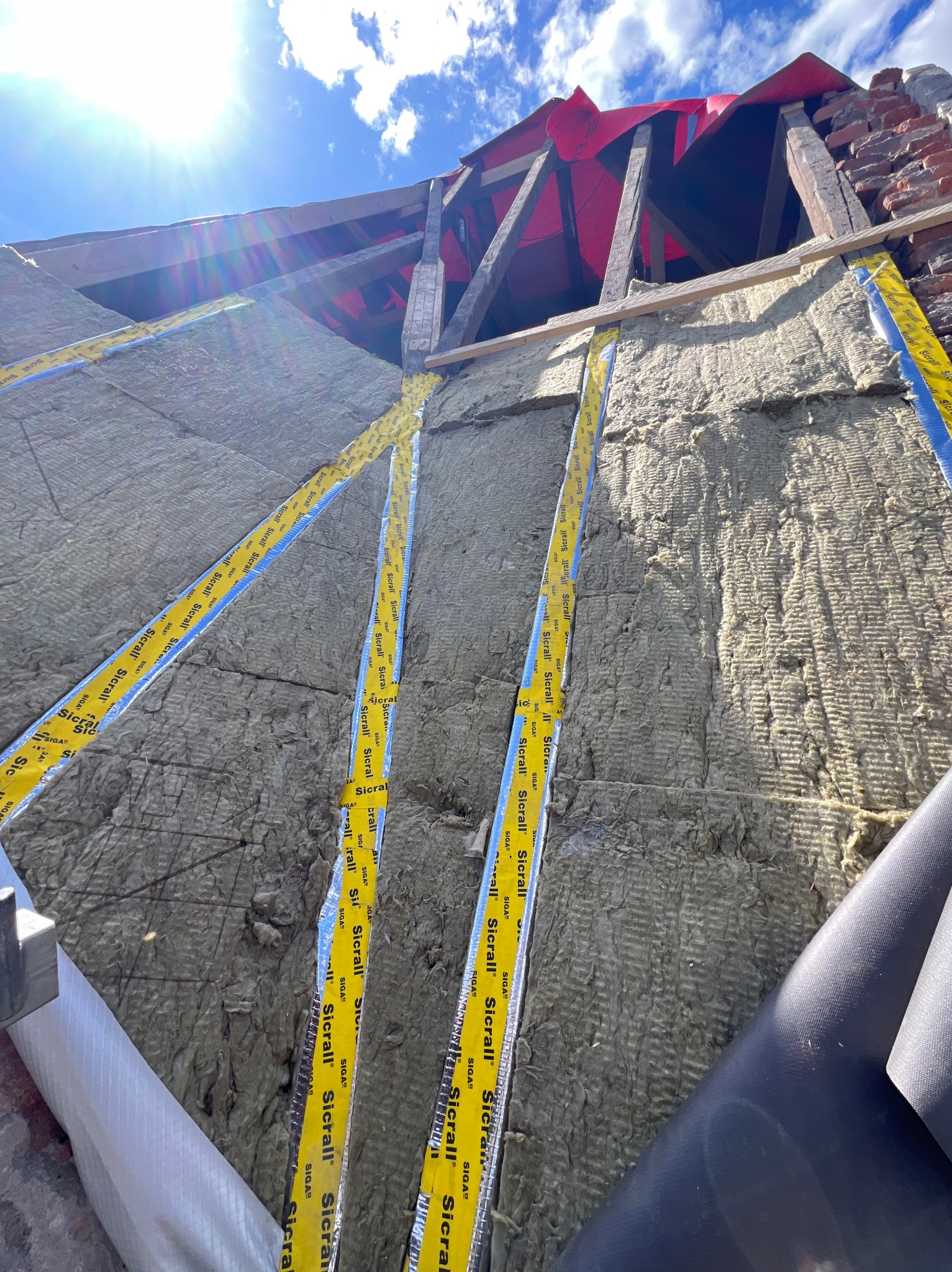 Ocieplenie poddasza wełną skalną z zewnątrz, zabezpieczone paroizolacją i taśmą SIGA. Widoczne drewniane elementy konstrukcji dachu i czerwona membrana dachowa.