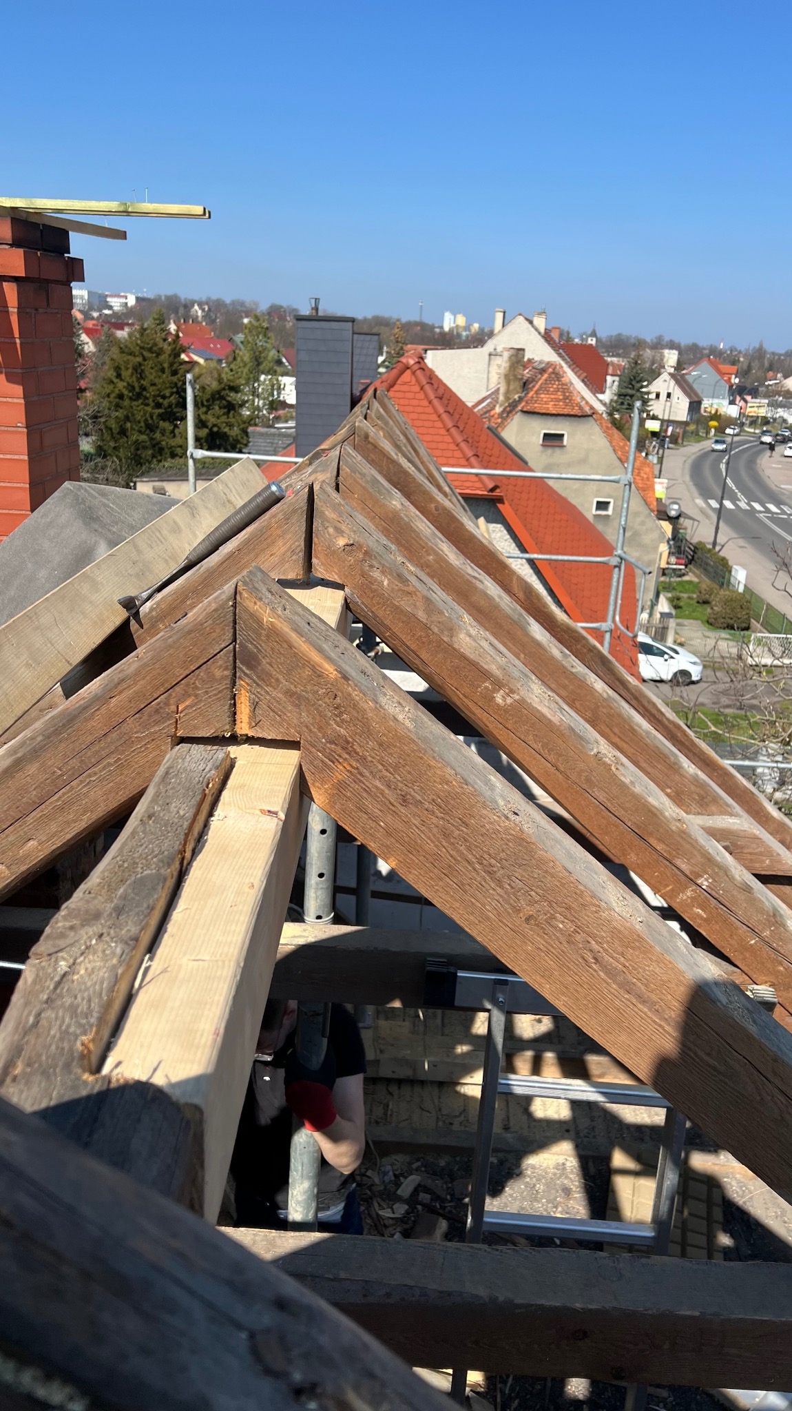 Konstrukcja więźby dachowej z drewnianymi belkami, widok z góry na tle zabudowy miejskiej w słoneczny dzień. Częściowo widoczna osoba w trakcie pracy.