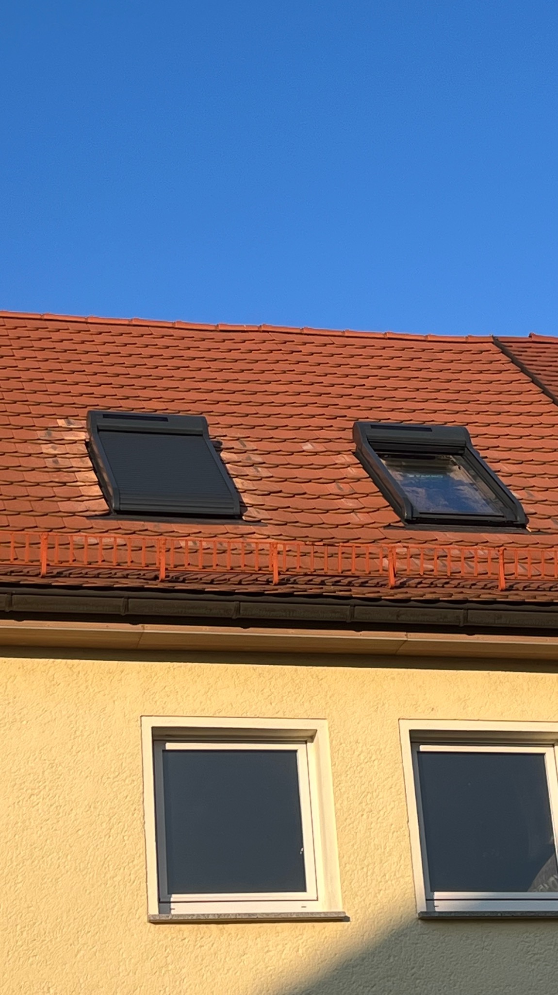 Czerwona dachówka z dwoma oknami dachowymi, jedno z roletą zewnętrzną. Fragment ściany budynku z dwoma oknami na dole kadru. Błękitne niebo w tle.
