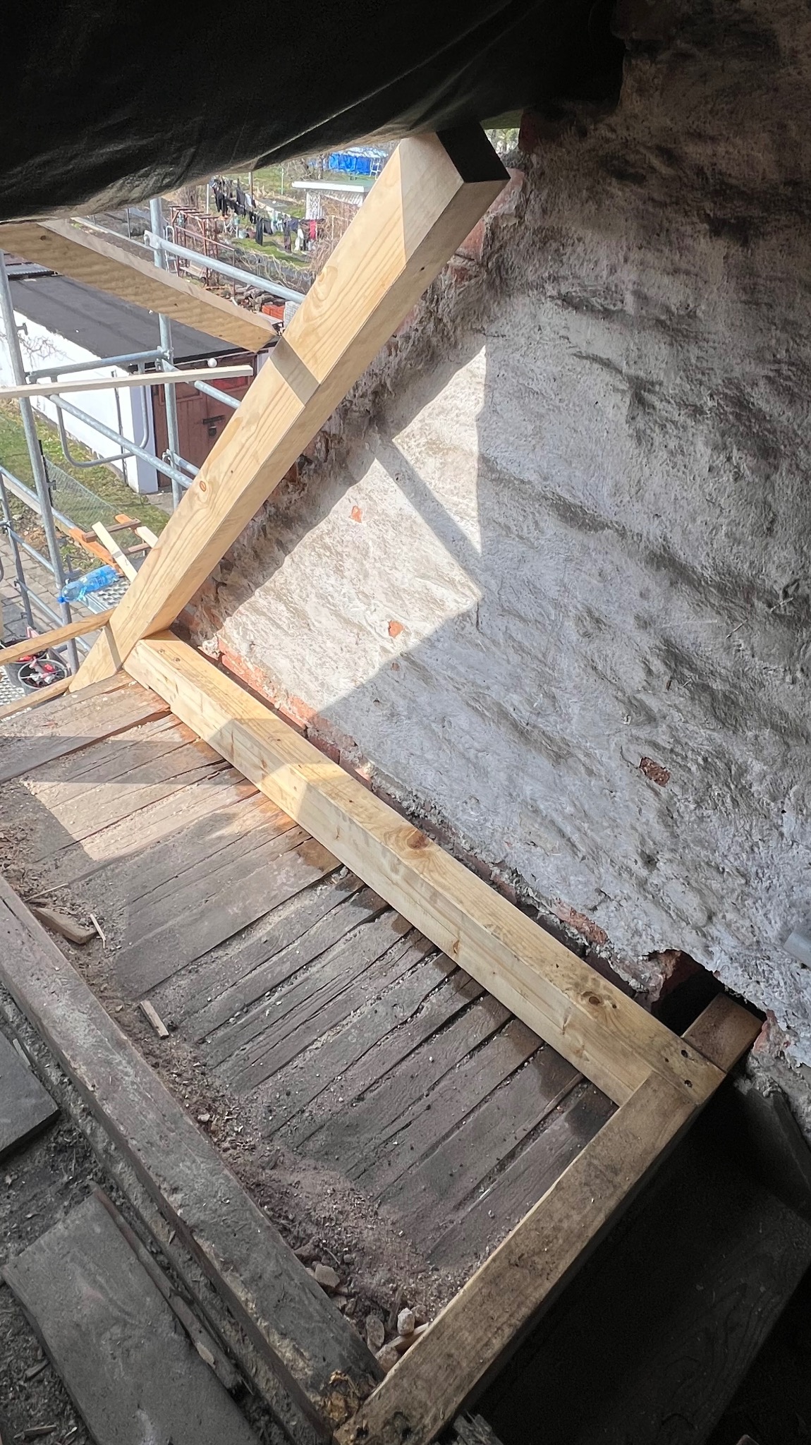 Konstrukcja drewniana dachu w trakcie budowy, widoczne elementy więźby, deski poszycia i fragment muru. W tle rusztowanie i zieleń.