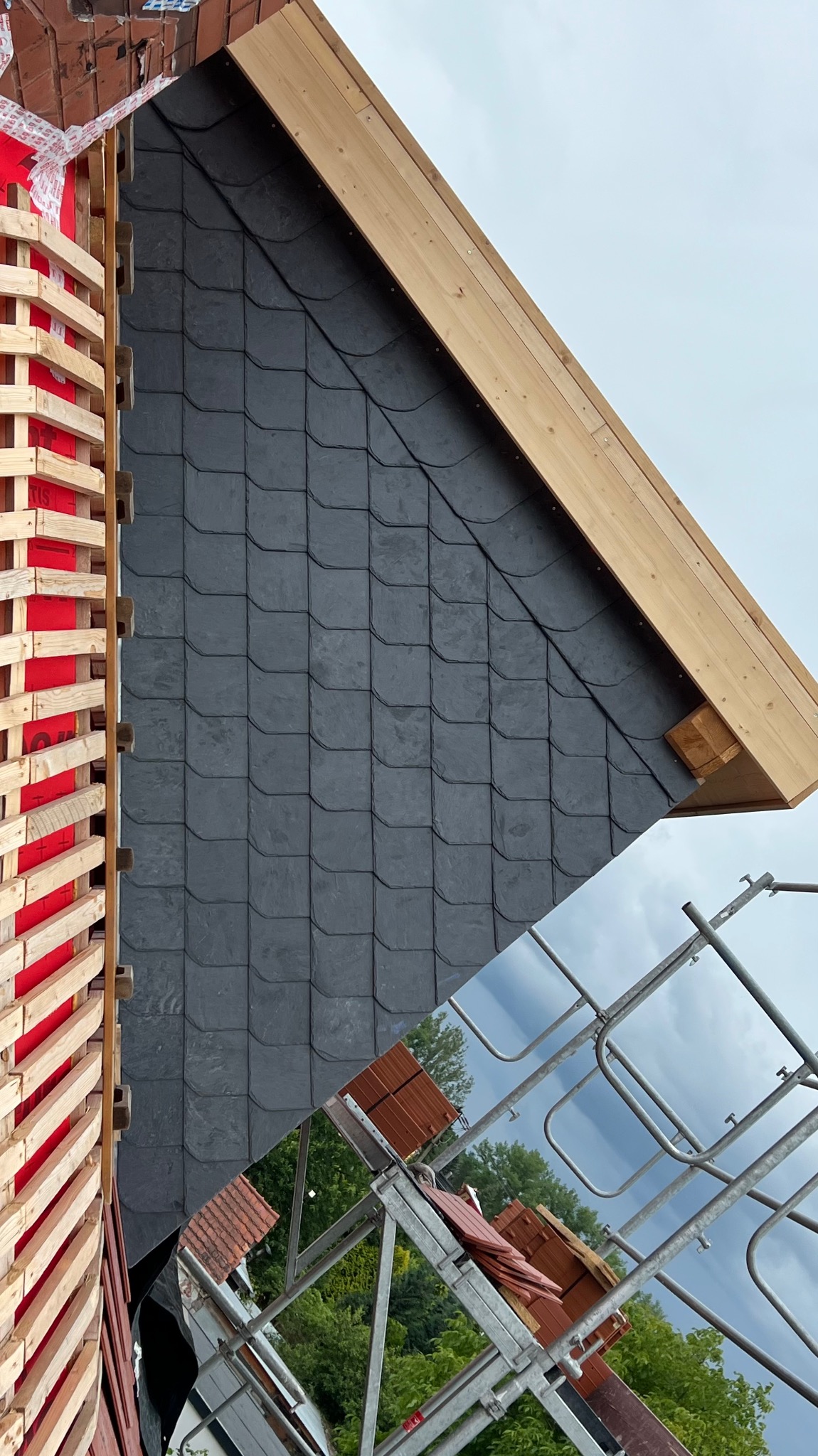 Fragment dachu pokrytego czarnym łupkiem, widok z góry. Widoczne drewniane elementy konstrukcyjne i rusztowanie z cegłami w tle.
