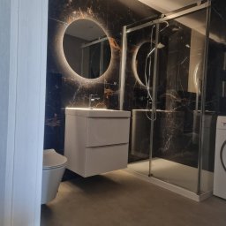 Mateusz Wardyński - Nowoczesna łazienka z czarnymi płytkami imitującymi marmur, okrągłym lustrem z podświetleniem LED, białą szafką pod umywalką i kabiną prysznicową.