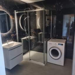 Mateusz Wardyński - Nowoczesna łazienka z czarnymi płytkami imitującymi marmur, kabiną prysznicową, białą umywalką z szafką i pralką. Okrągłe lustro z podświetleniem.